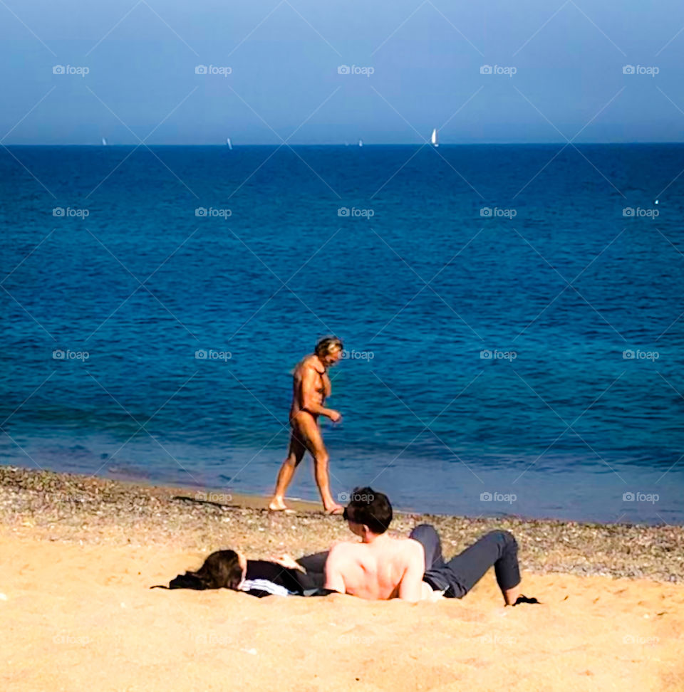 Nudist with great tan, strolls along the beach past 2 sunbathers in Barcelona. The Mediterranean Sea is deep blue and the sails of yachts can be made out on the horizon 