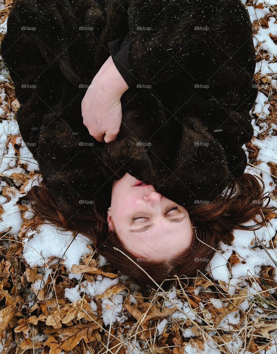 portrait of a light-skinned girl lying among the snow and leaves in a black fur coat