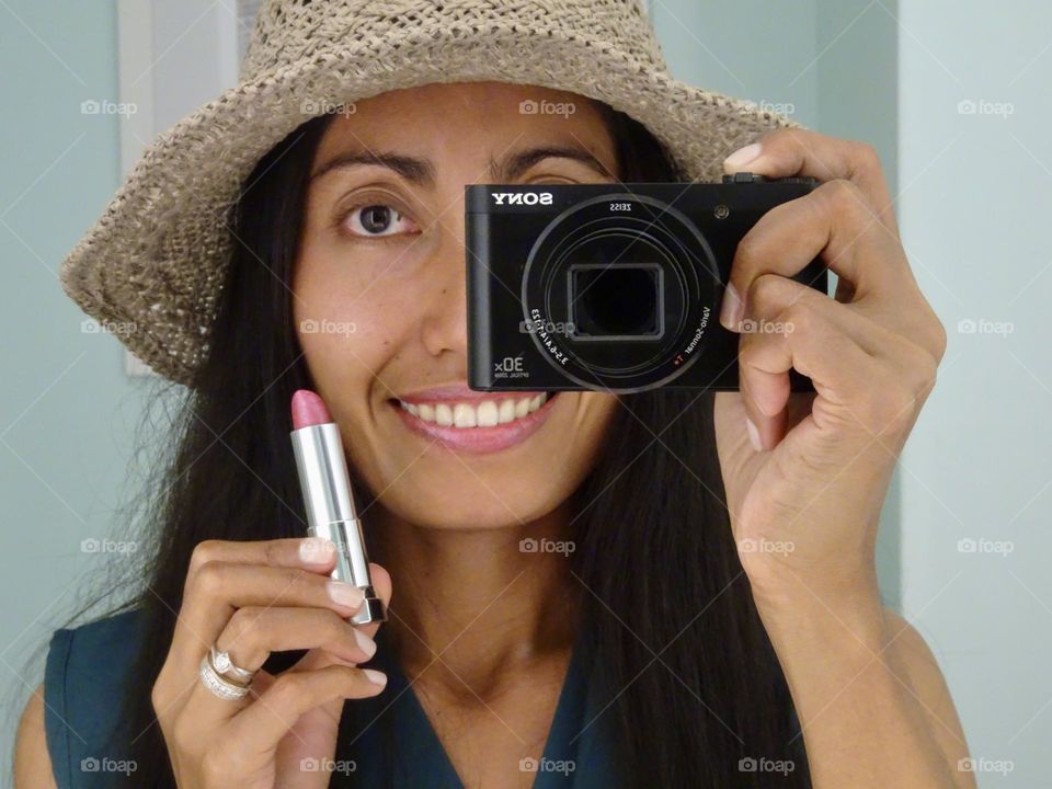 Close up reflection of smiling woman holding a camera and lipstick and wearing gray hat.