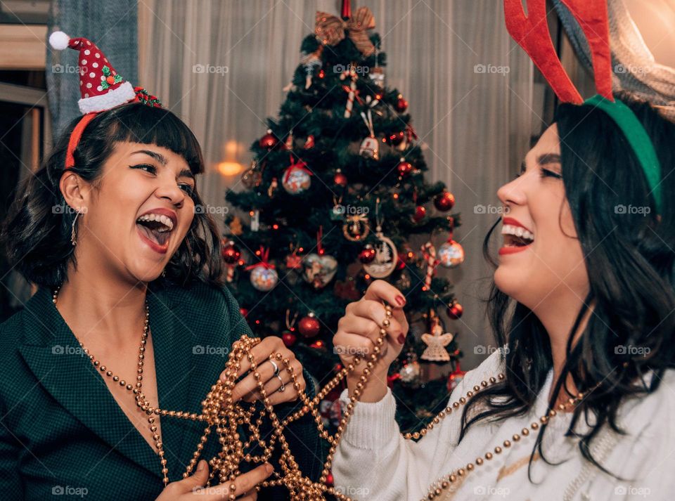 Two young female friends laughing and having fun while decorating a christmas tree at home