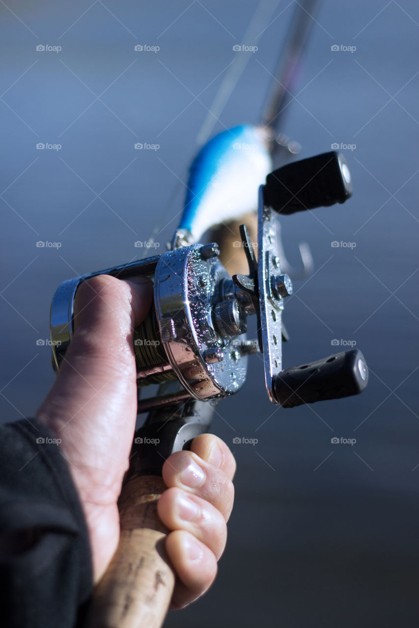 Male hand on a spool of a round fishing reel attached to a fishing rod with fishing lure and blurred nature background.