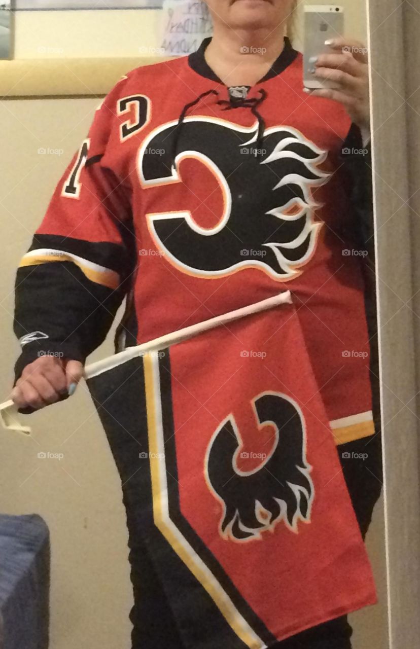 Middle aged, plus size woman is wearing her Funky, colourful, red, black, yellow and white, Calgary Flames NHL jersey, and holding in her hand a matching flag to the hockey game tonight