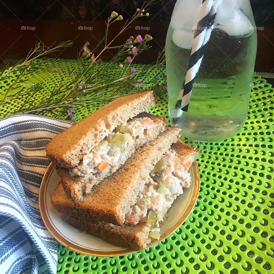 A yummy and toasted tunafish salad sandwich on a lime green placemat and a bottle of cold ice water with black and white straws, a blue and white striped napkin and lavender flowers for decor.