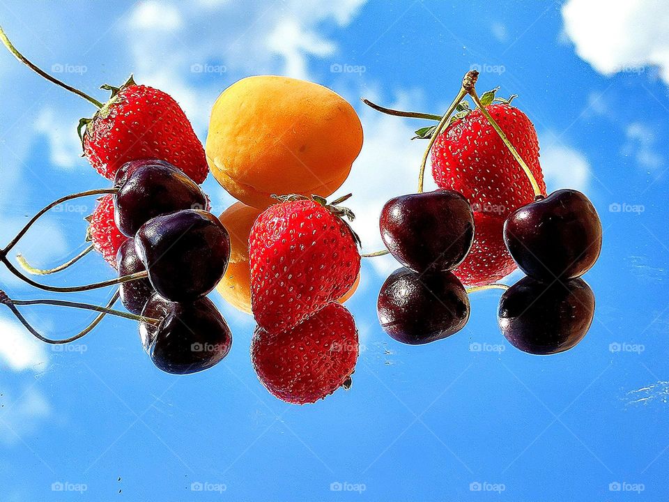 Summer.  Time for fruits and berries.  On the mirror surface lie: yellow apricot, red strawberries and dark burgundy cherries.  Blue sky reflection with white clouds