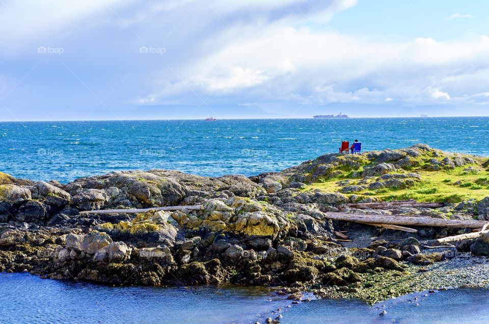 A distant view of two people sitting in wooden chairs watching and relaxing in a scenic seaside setting