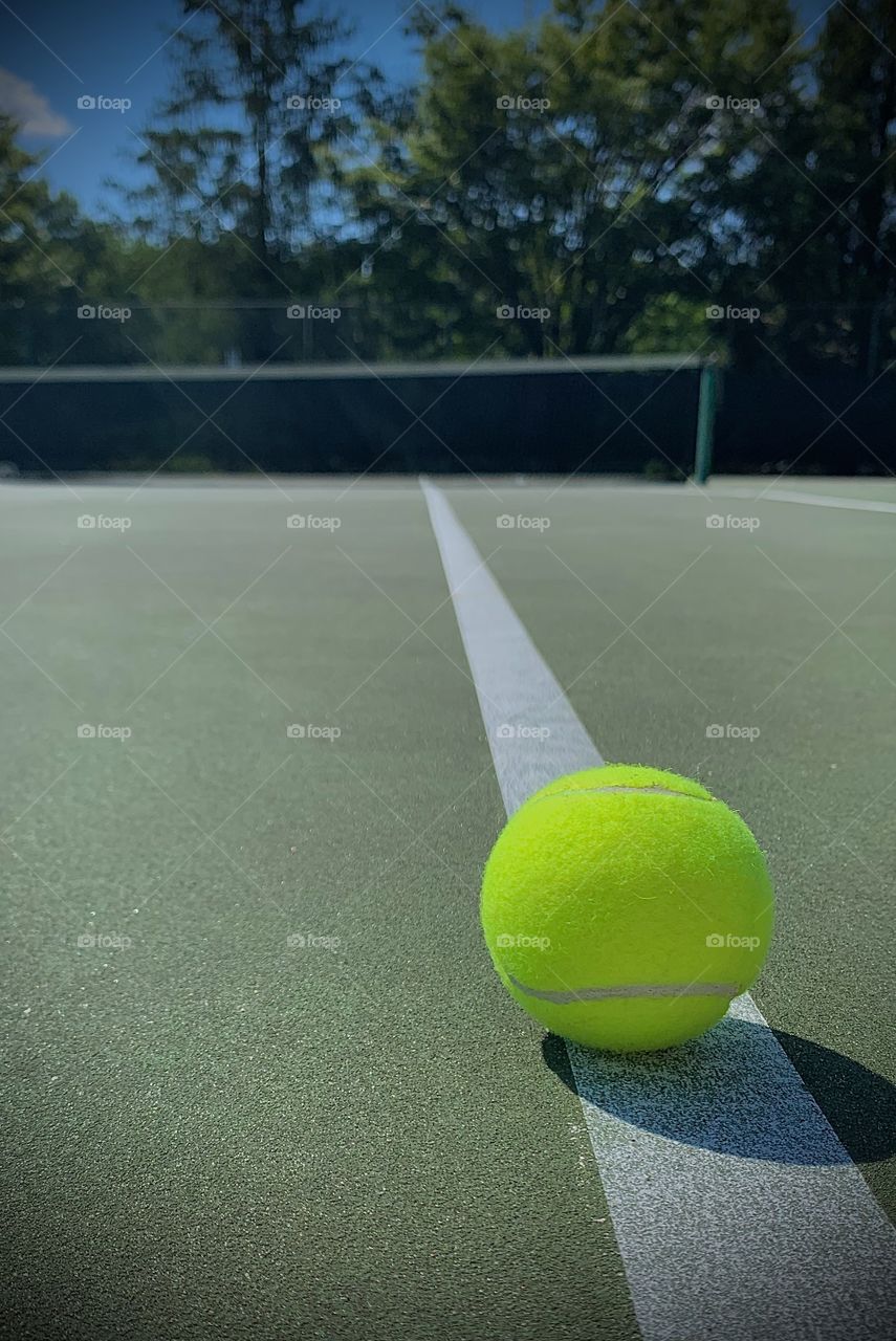 Colorful photo of a tennis ball on the line with a shadow. Looking out onto the court and net.  