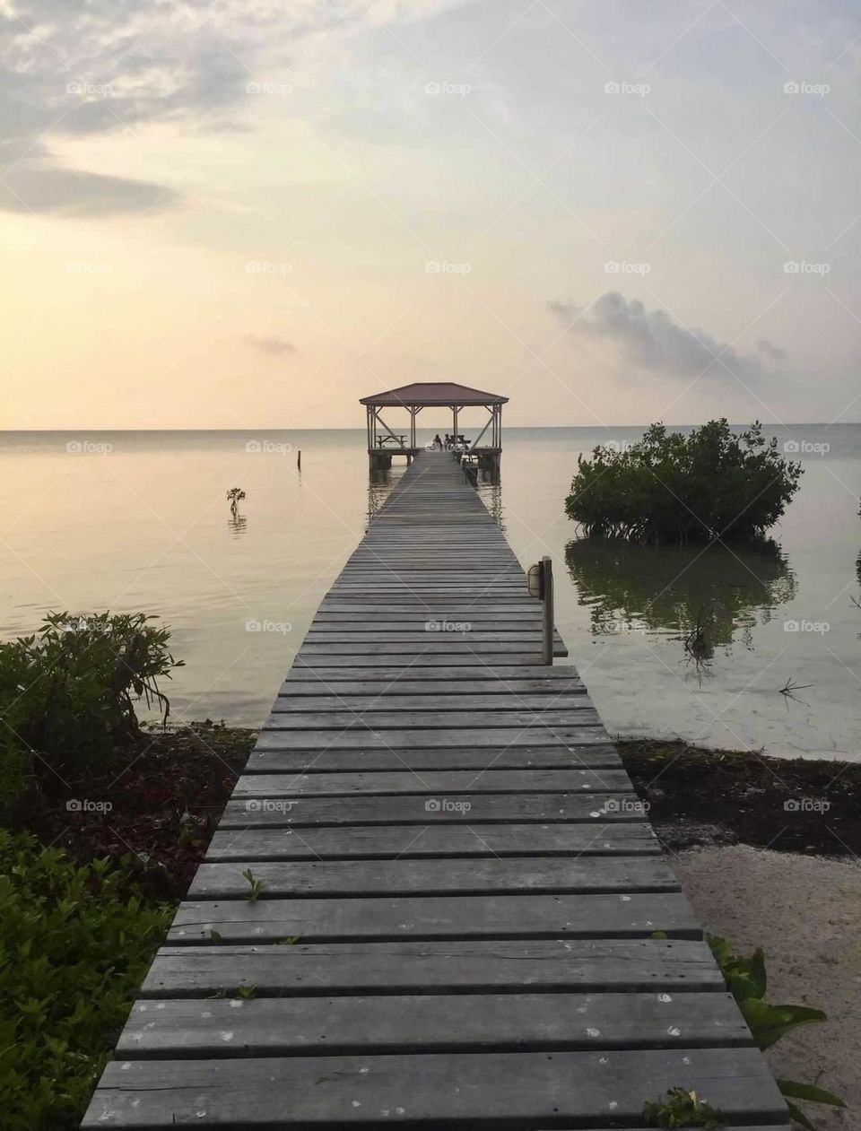 A wooden walkway leads to the end of the dock. A gazebo overlooks the lake with the sun setting in the sky. Sand and green trees create an inviting environment. 