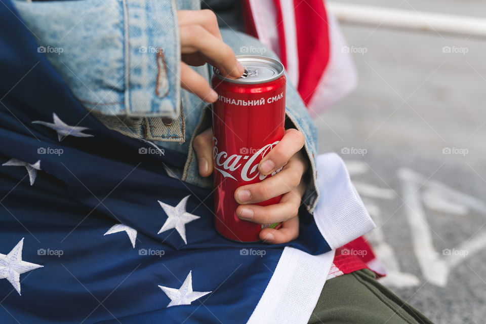 a guy sits on the street, holds a can of Coca-Cola in his hands, opens it.  the guy is dressed in denim clothes.