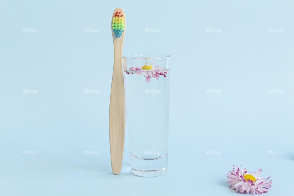 An ecological, wooden and reusable toothbrush with multi-colored bristles stands with a glass of water and a summer daisy, on a blue background.