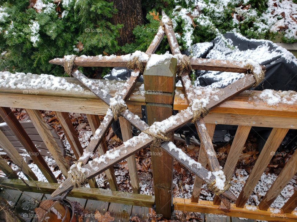 A rustic wooden star is seen hanging on a wooden rail with a light freshl amount of snow