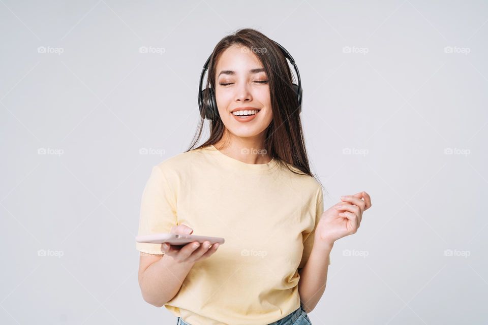 Emotional portrait of Young happy asian woman with long hair in yellow shirt and jeans listening music by headphones on grey background