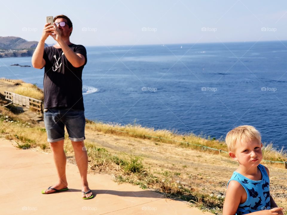 father with mobile phone and a grumpy son. Ocean in background