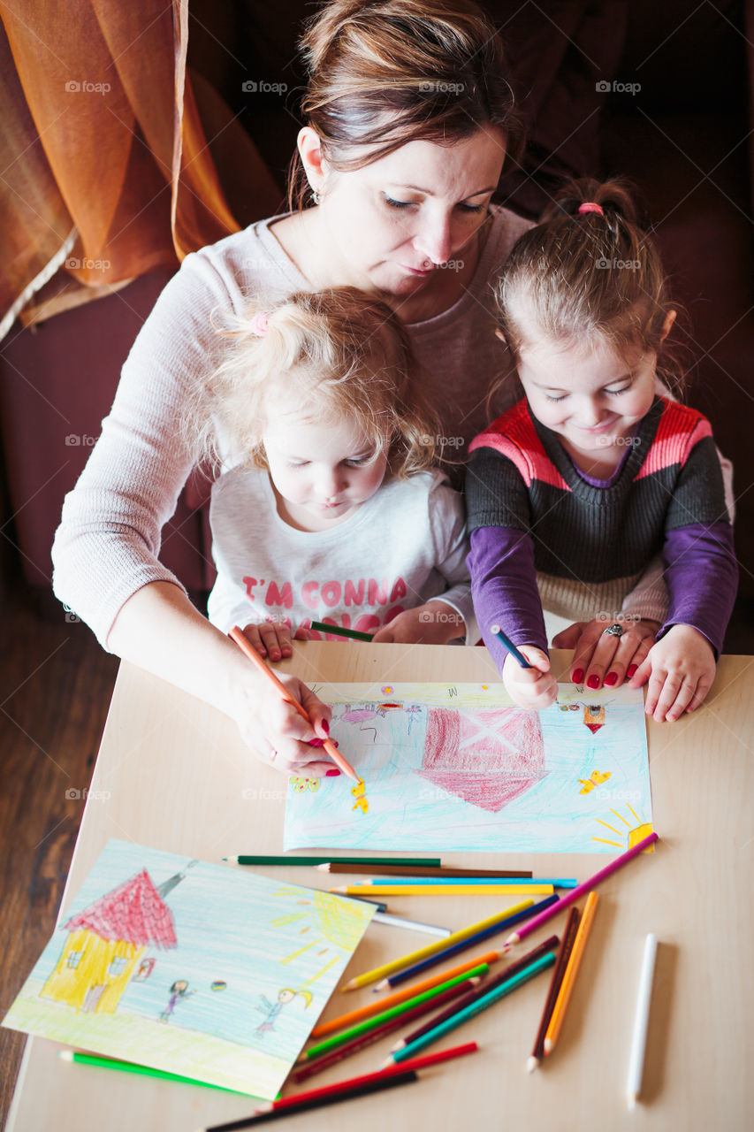 Mom with little girls drawing a colorful pictures of house and playing children using pencil crayons sitting at table indoors. Shot from above