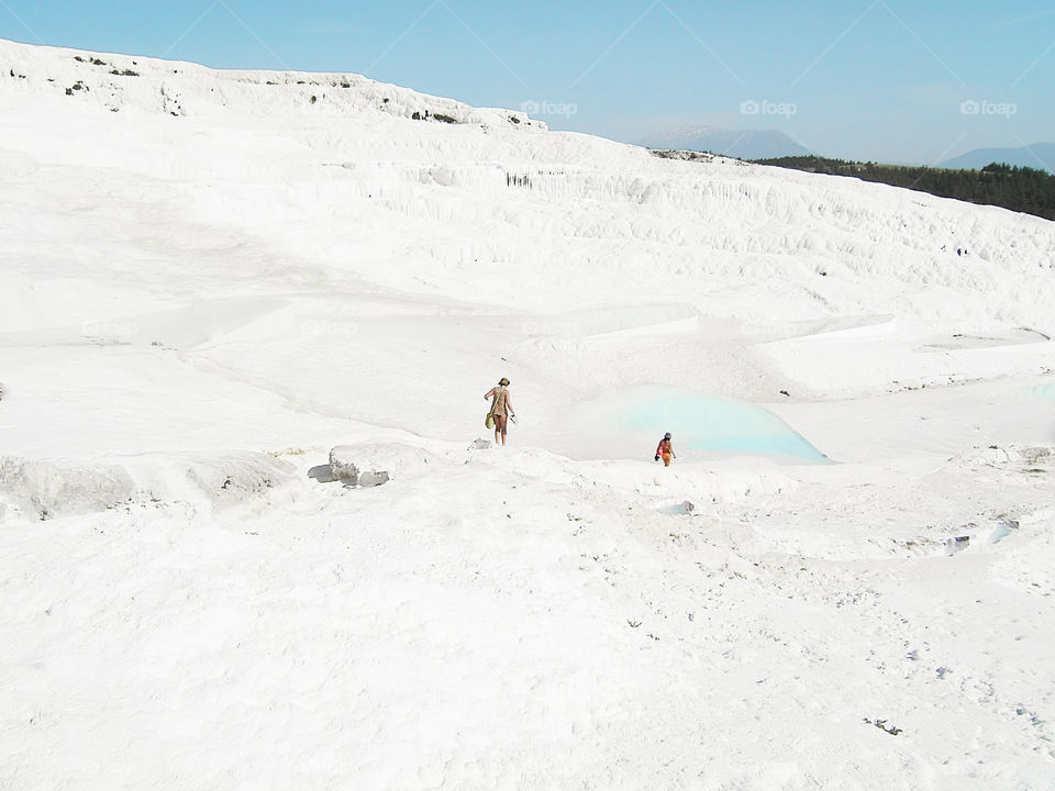 Tiny humans walking by the white chalk mountain with blue lagoons in Pamukkale 