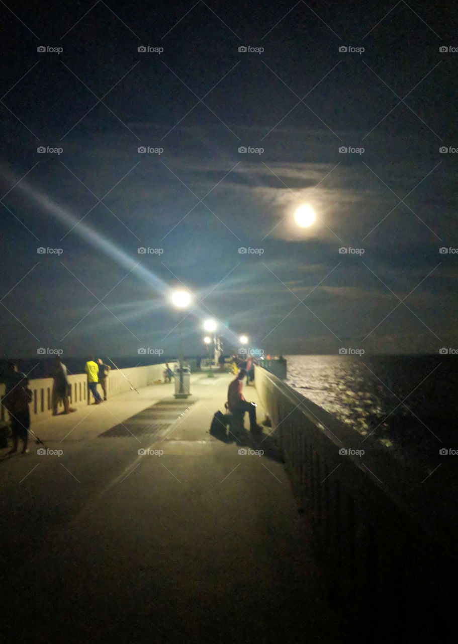 The moon shines bright on this clear night in the fishing pier. Fisherman can be seen waiting for their next catch. 