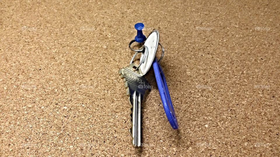key hanging. This is a shot of a key on the board on a notice board with a pin.  
a little blue pin with a blue key holder.