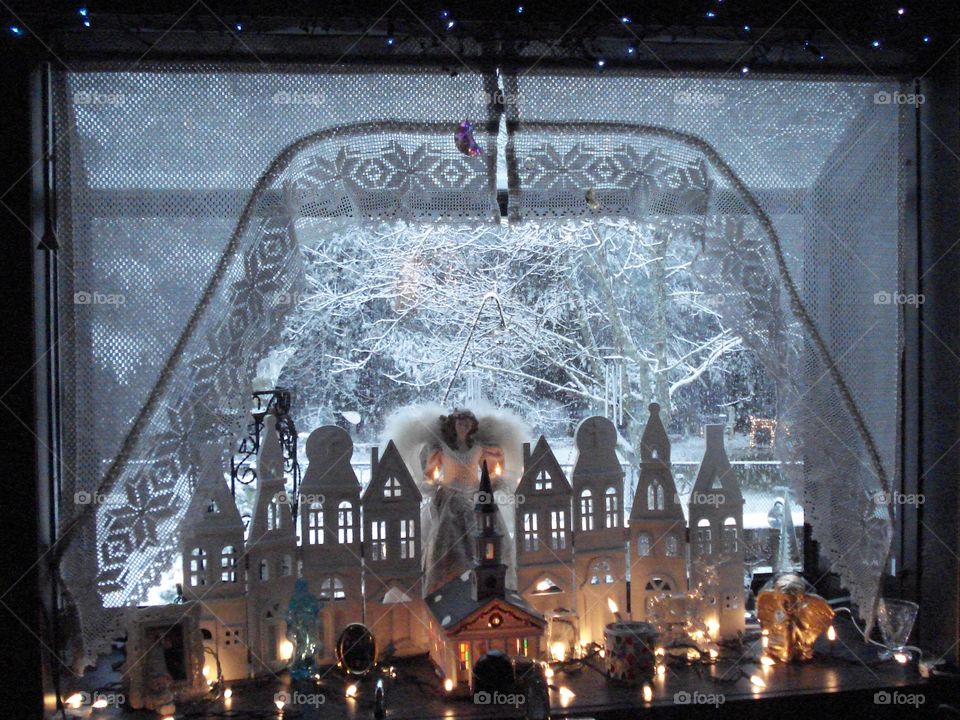 Winter is coming, snowstorm seen through window! Bay window decorated for winter with Angel and lights. Town silhouette in background.❄