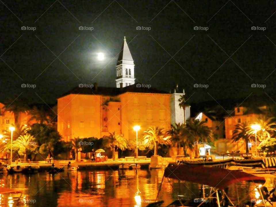 Golden lit Italianate buildings reflected in a Croatian harbour with a full moon reflecting on a white stone Venetian tower