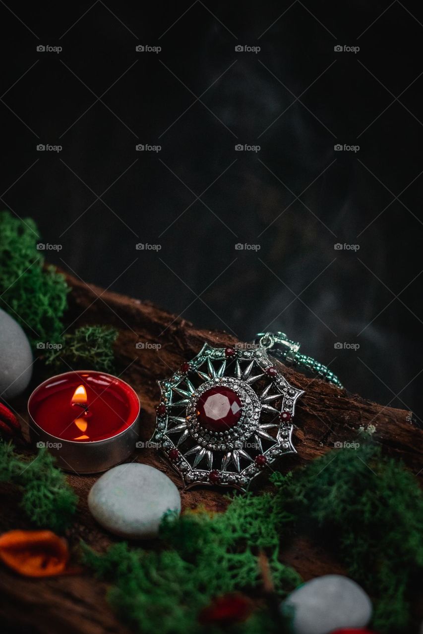 One ancient amulet with red stones on a silver chain lies in a natural tree bark with green moss, gray stones, a burning candle and a blurred jet of smoke on a black background, close-up side view. The concept of vintage jewelry, ancient amulet, esot