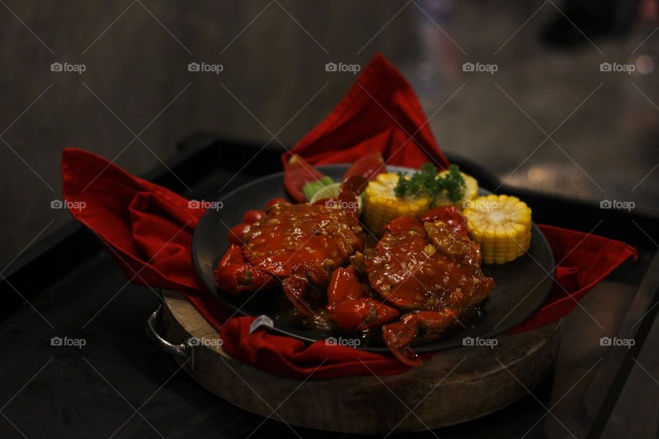 Crab flavored in creamy garlic sauce served with corn, tomatoes, lemon wedges and cherries on a black plate set on a red cloth set in a wooden setting. Asian flavor crab.