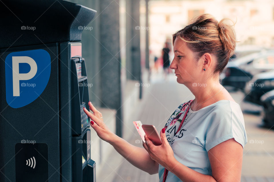 Woman buying a ticket for parking using debit card or mobile phone, standing next to parking machine