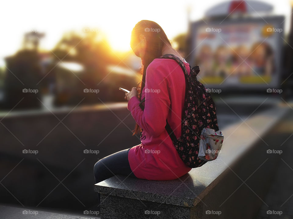 Young woman student with a backpack using mobile phone during the golden hour in the city 