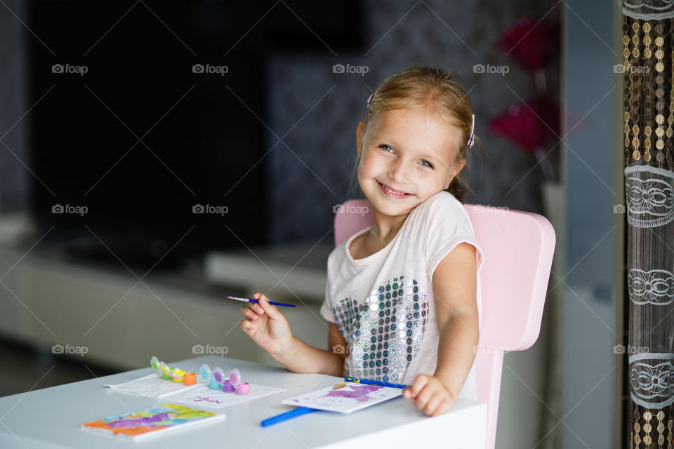 Little Caucasian girl with blonde hair painting at home. She is using table and chair from IKEA