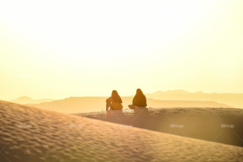 Two young friends sit together and watch the sunrise on top of a sand dune in the desert 