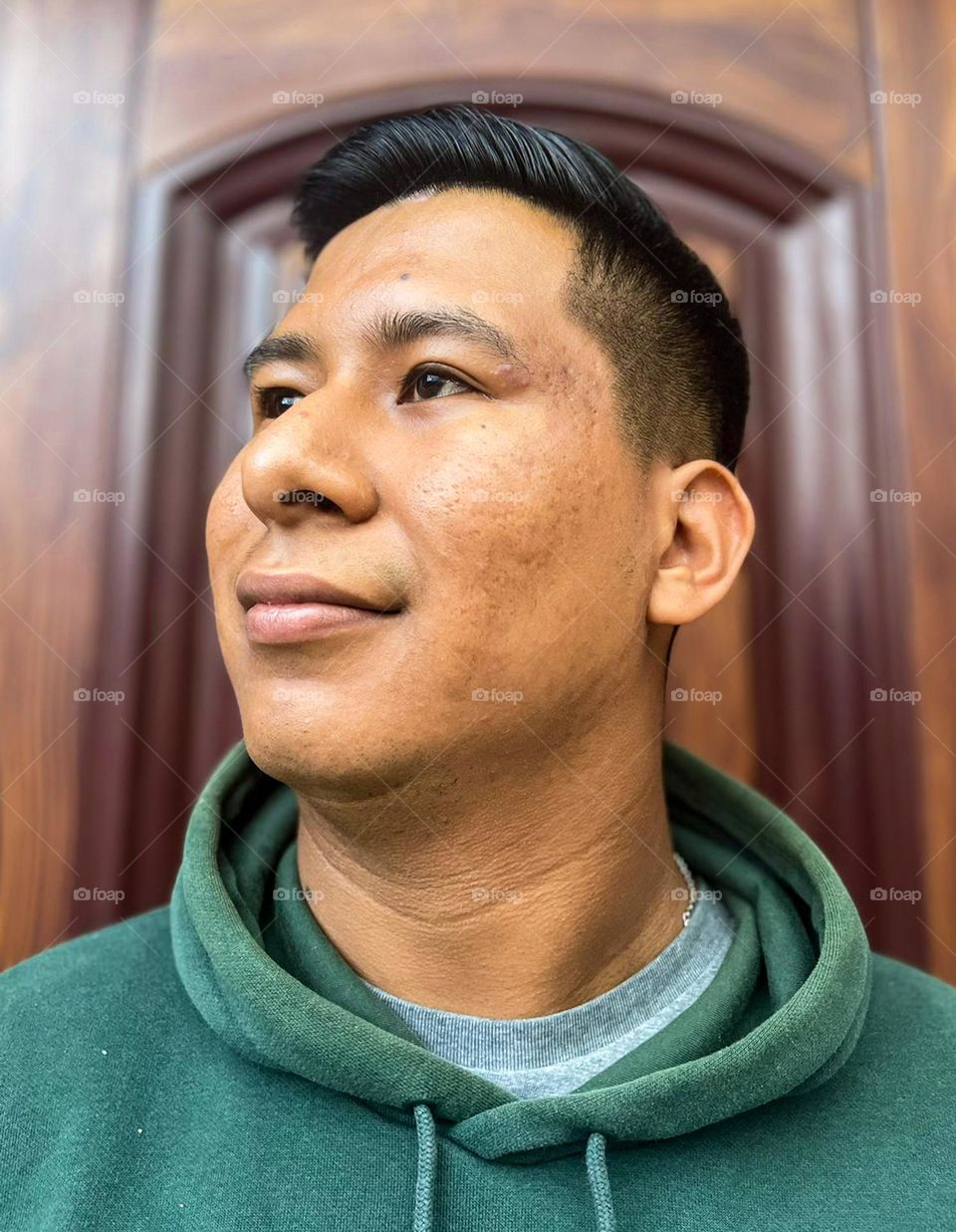 Portrait dark-skinned man, with acne and acne scars, wearing a green sweater and standing with a wooden door with blurred background.