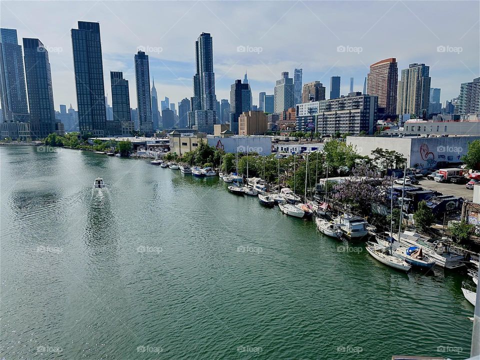 This is “Newtown Creek” with its boats on the water as well as on the shore seen from the “Pulaski Bridge” that connects LIC to “Greenpoint”, Bklyn. Across the “East River” we see “Manhattan” incl. the “Empire State Bldg”. 2024. Hypnotic Productions