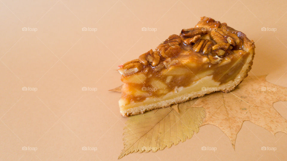 Useful baking. An appetizing piece of apple pie with pecans on autumn leaves on a plain light brown background. Autumn inspiration