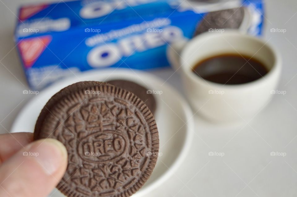 A persons fingers holding an Oreo cookie and a cup of coffee in the background