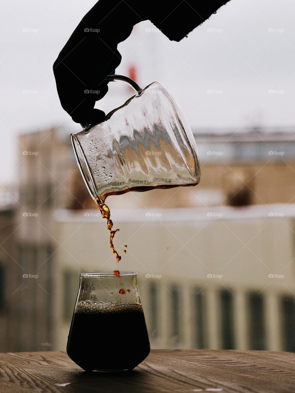 An unrecognisable man's hand pours fresh, hot coffee into a glass cup placed on a wooden table near the window, still life.