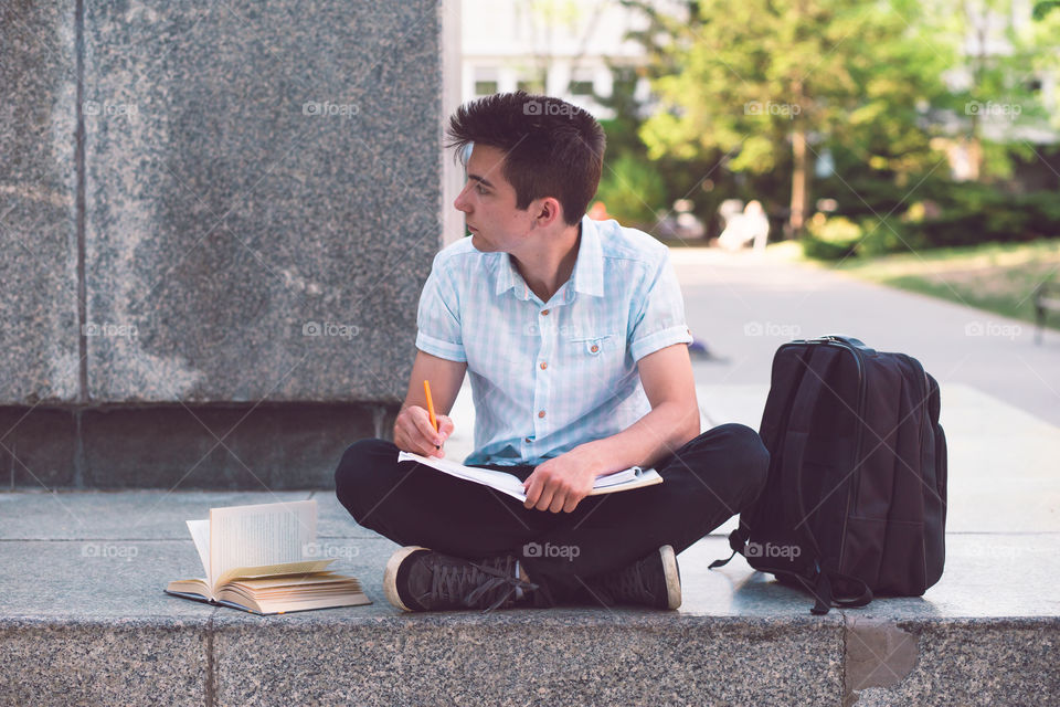 Student making the notes learning from books sitting on a monument outside of university. Young boy wearing a blue shirt and dark jeans