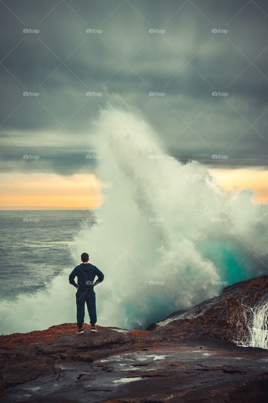 Brave Man standing in front of a big wave crushing on the rocks with huge power at sunset 