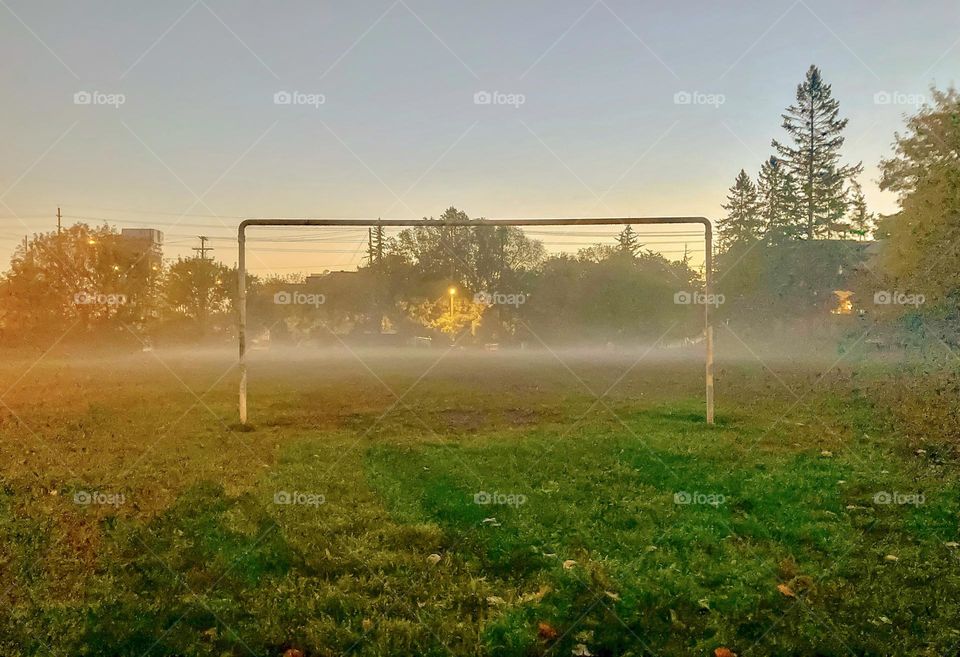 Through the net at an empty football pitch draped in morning mist.