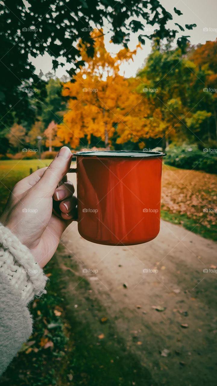 The hand of a young Caucasian girl holds a metal mug on a background of an orange tree in an autumn park on a gloomy evening, close-up side view.