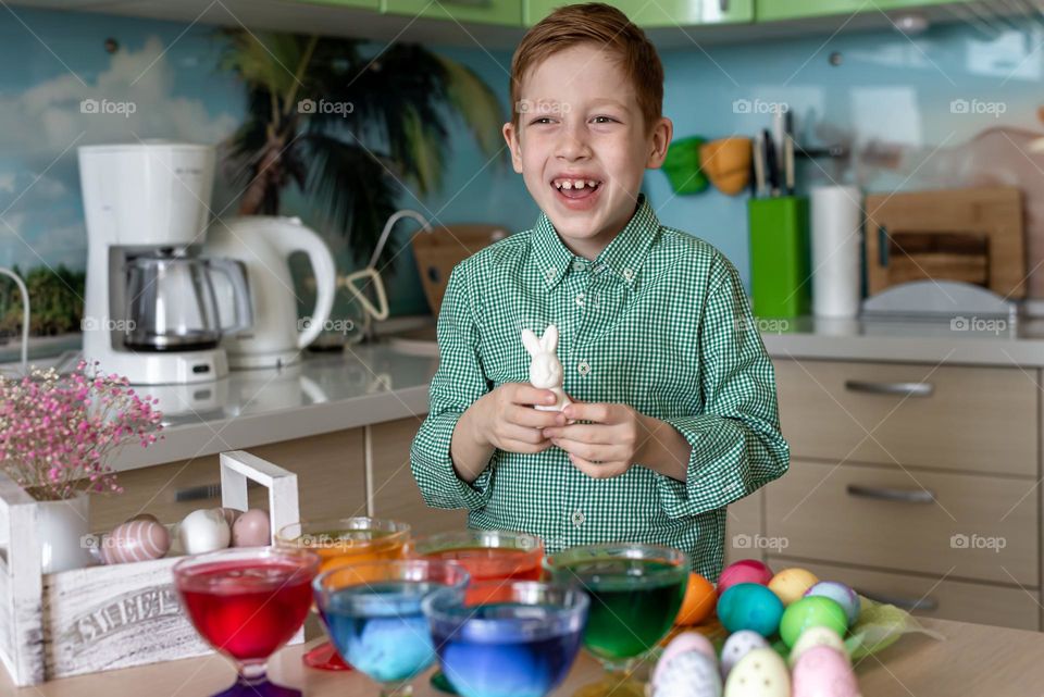 A red-haired boy child in a plaid shirt paints Easter eggs in the kitchen at home and laughs. Cooked with my own hands