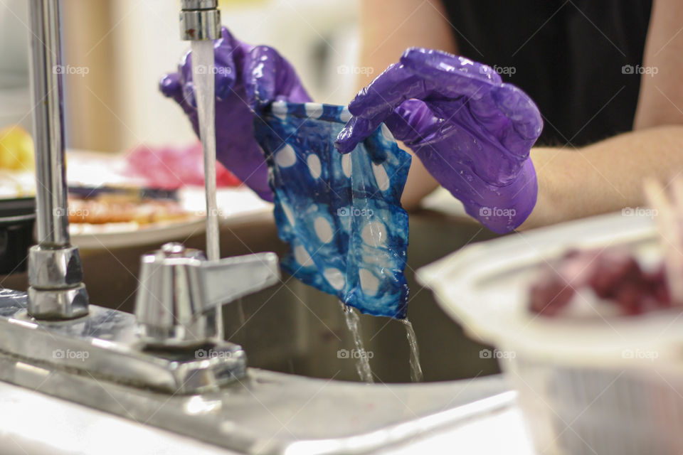 Gloved hands holding a hand dyed piece of fabric under running water in a sink 