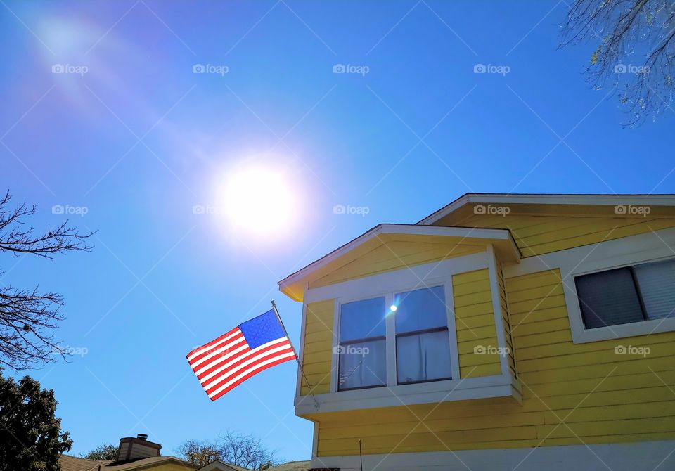 USA flag on a yellow residential home on a clear blues blue sky day and the sun out.