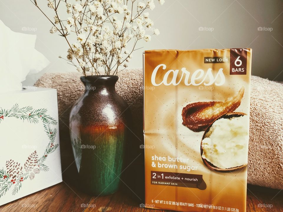 A six pack of the new look Caress shea butter and brown sugar bar soap on a wood surface next to a vase with small white flowers and a tissue box.