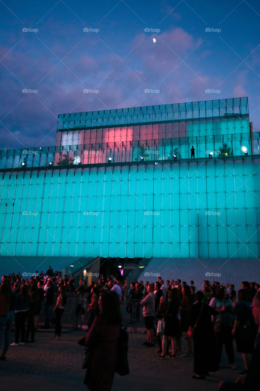 Festival event party concert with people standing on a square in a city center at night. Backlit glass construction above them