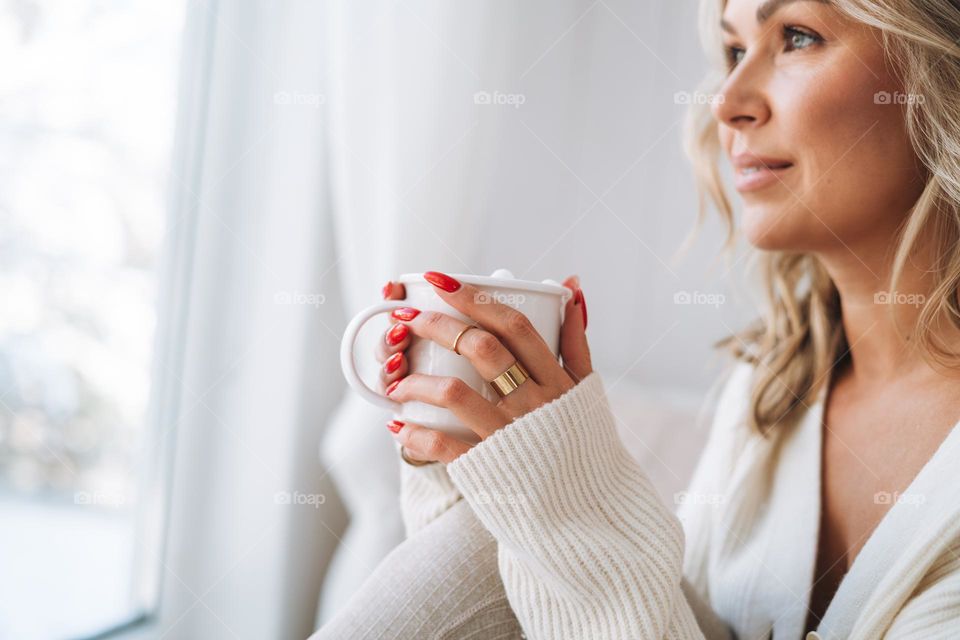 Blonde woman with red manicure holding cup with cocoa marshmallow at home in winter season 