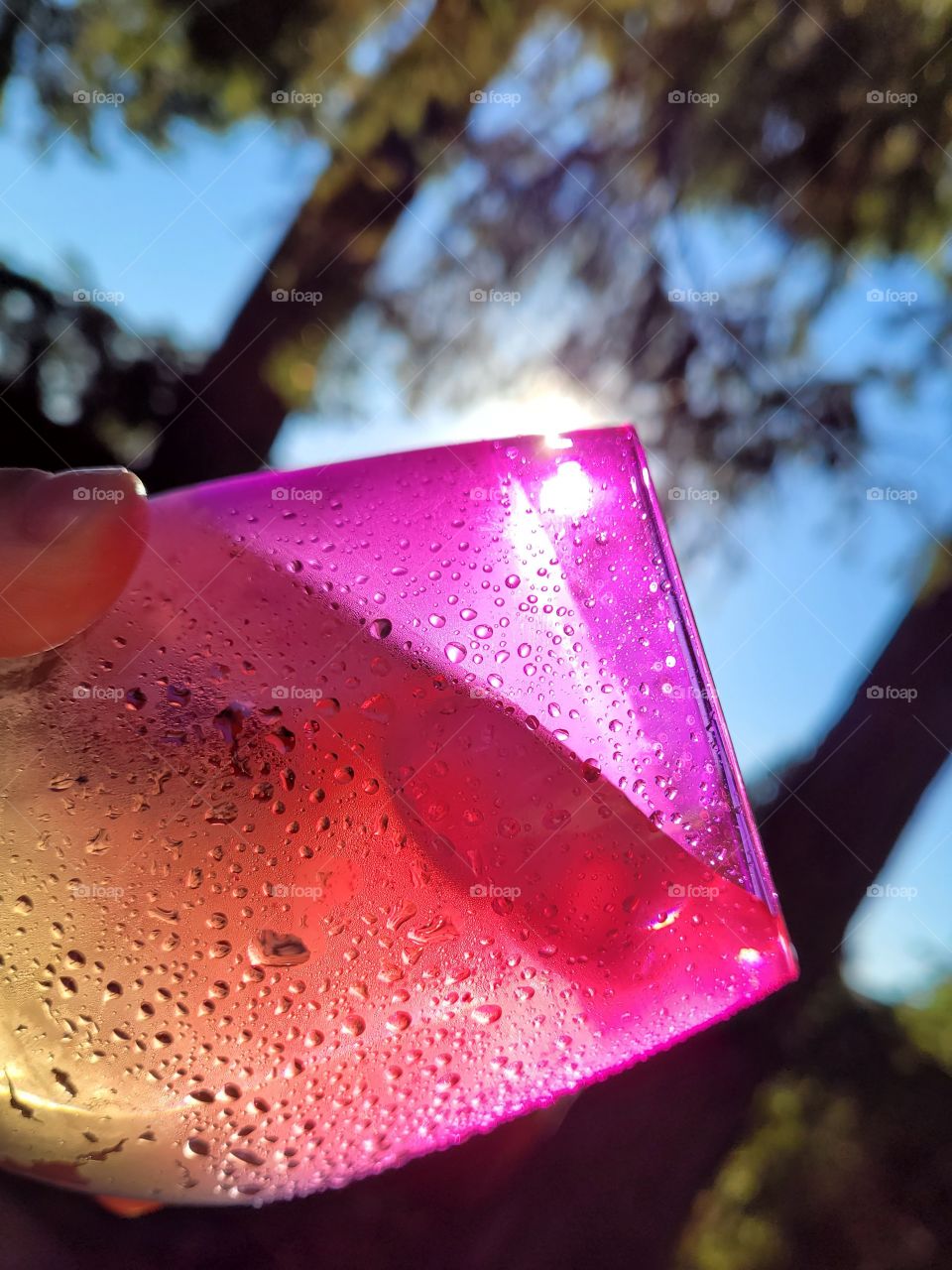 hand holding neon multi colored sunset/sunrise glass with pinks reds and yellows, cold drink with ice and condensation drips, angled up to see bright sun, blue sky, and Summer trees