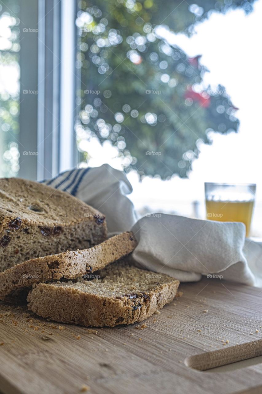 The fresh morning light enters the kitchen where there is a cereal bread and raisins on a wooden board with a linen towel and orange juice for breakfast 