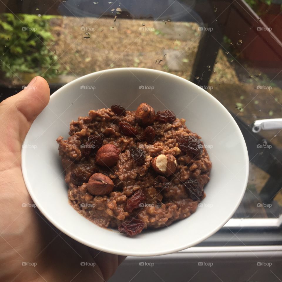 Chocolate flavoured oats in a white bowl with hazelnut nuts and raisins held by a hand 
