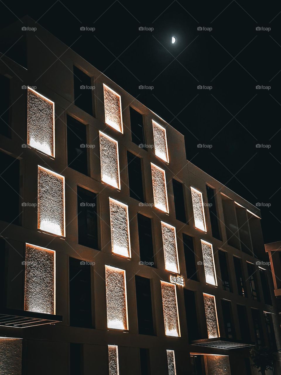 A building with lit up rectangular windows at night with the moon in the sky.