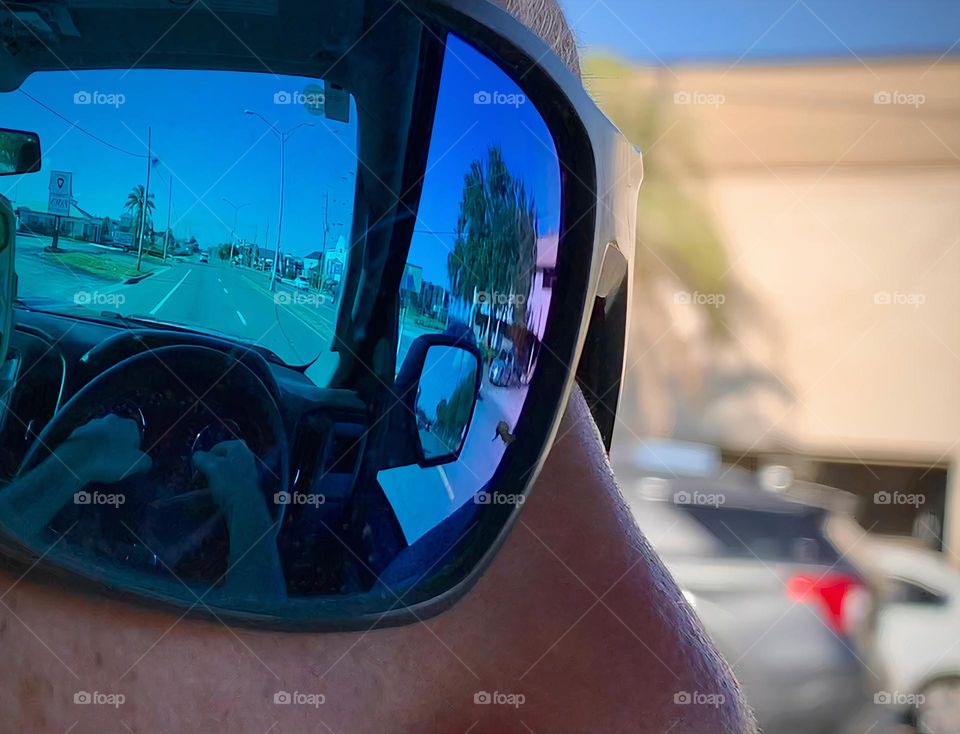 Commuting: driver man driving with sunglasses passing vehicles seen from the left window and reflection of the road and the steering wheel with his hands holding it, the interior of the pickup truck and the side mirror from his glasses.