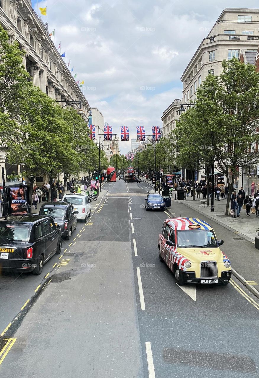 Multicolored taxi and other cars, buses and bikes in the background on crowded street with british flags and trees.