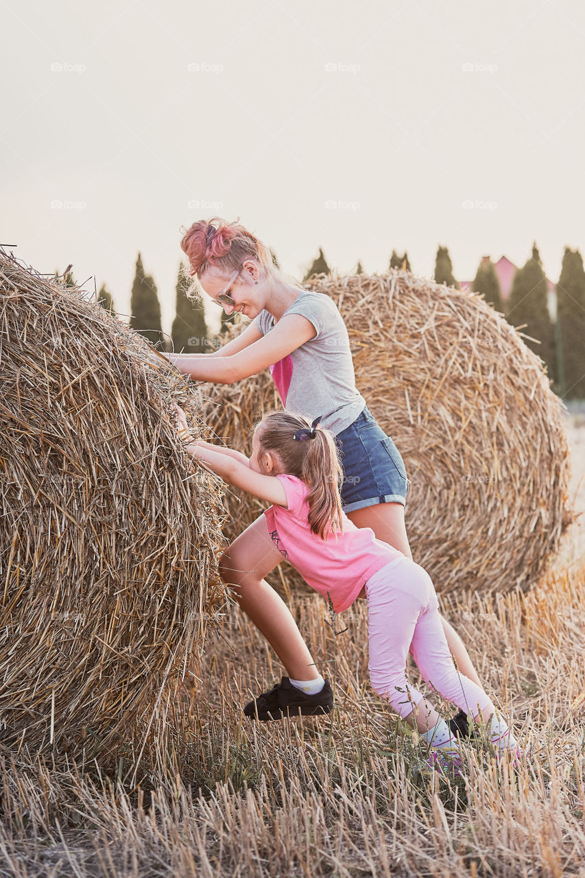 Sisters, teenage girl and her younger sister playing together with hay bale outdoors in the field in the countryside. Candid people, real moments, authentic situations